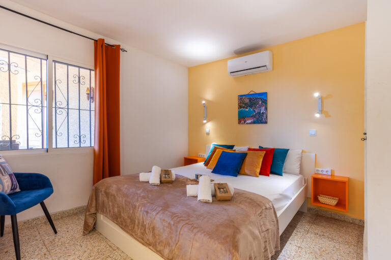 Habitación con cama doble, cojines coloridos, pared amarilla, cortinas naranjas, silla azul y aire acondicionado.