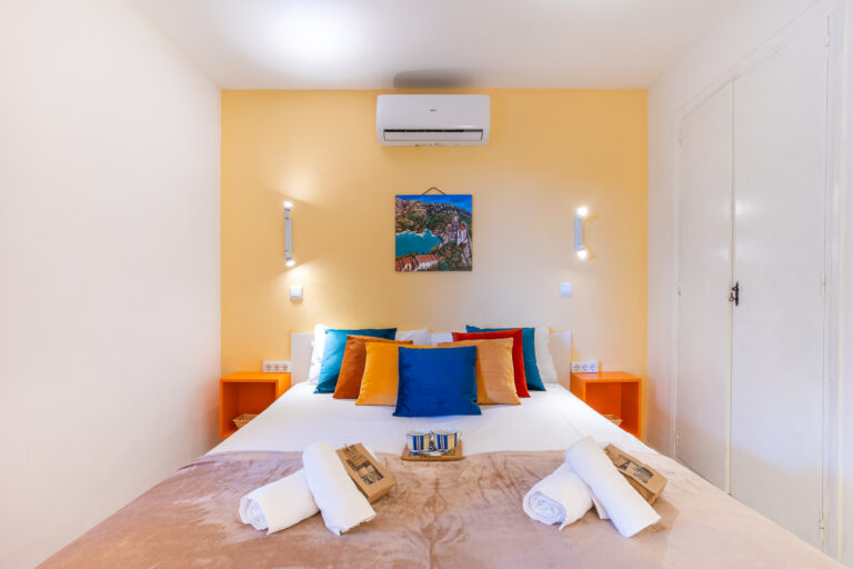 Habitación luminosa con cama doble, cojines coloridos, pared amarilla, aire acondicionado y arte enmarcado sobre la cama.