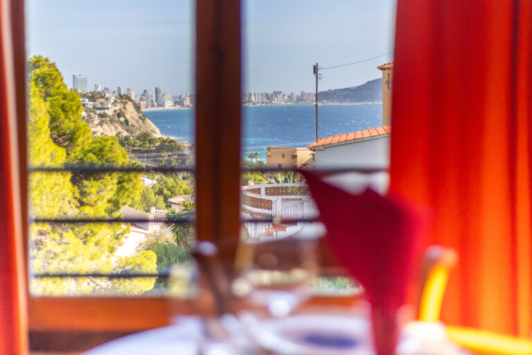 Vista desde una ventana de un paisaje costero, mar azul, acantilados verdes y ciudad con rascacielos al fondo.