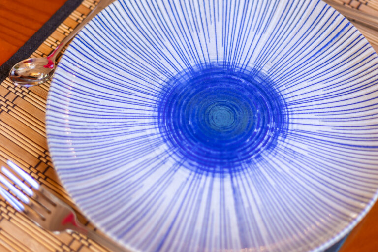 Platillo con diseño de líneas azules radiales sobre mantel de bambú, acompañado de tenedor y cuchara metálicos.