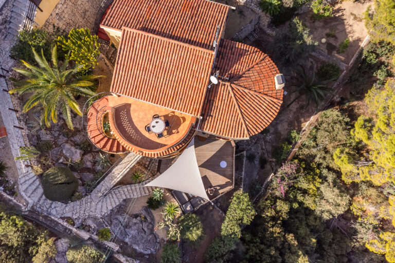 Vista aérea de casa con techo de tejas rojas, jardín con palmeras, mesa en terraza y sendero de piedra. Vegetación alrededor.