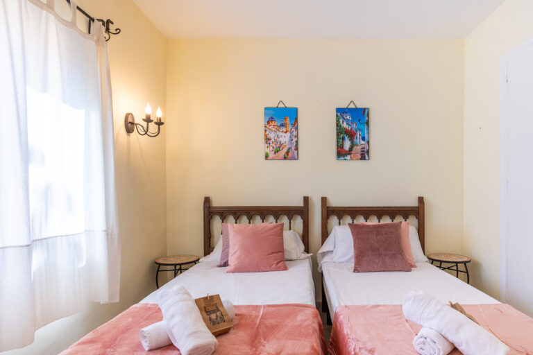 Habitación con dos camas individuales, cabeceras de madera, cojines rosados, lámpara de pared y cuadros coloridos en la pared.