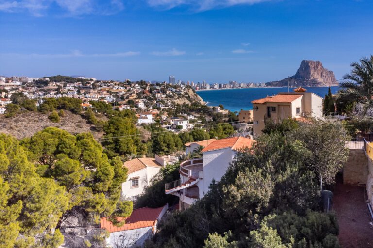 Vista panorámica con casas blancas, montañas, vegetación verde, mar azul y un peñón prominente en el horizonte.