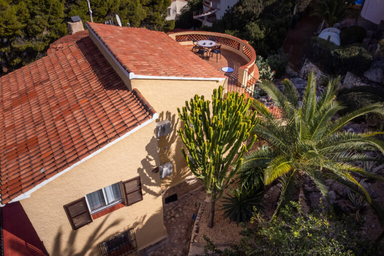 Casa con tejado de tejas rojas, terraza redonda, cactus grande y palmera. Jardín con plantas verdes y camino de piedra.