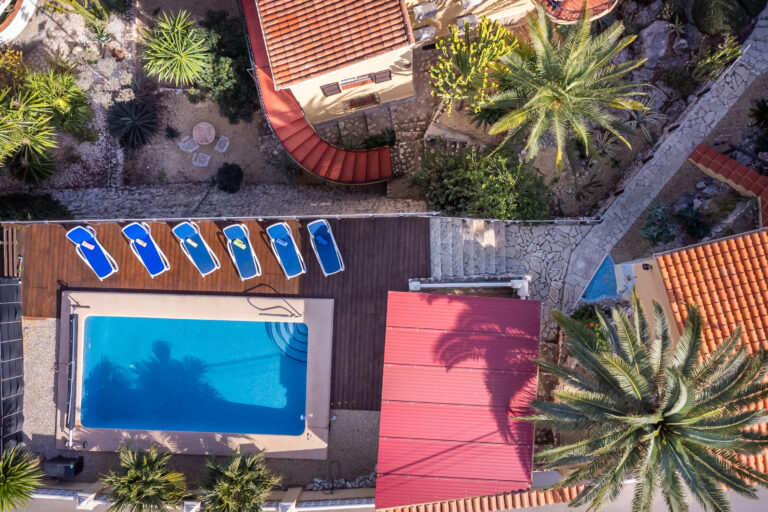 Vista aérea de piscina rectangular con seis tumbonas azules, rodeada de palmeras y senderos de piedra en jardín.