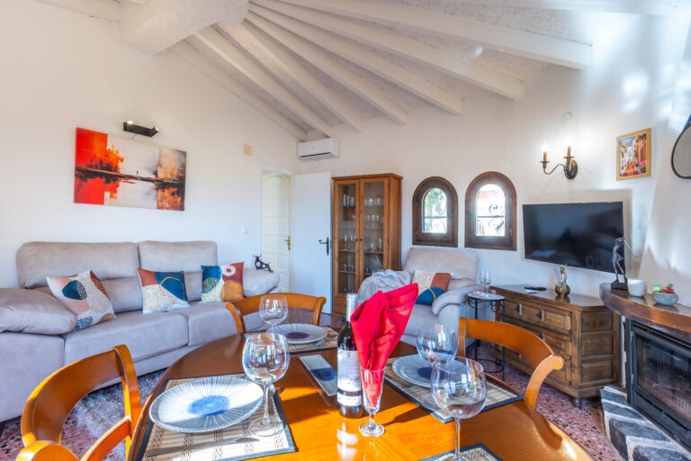 Sala acogedora con techo abovedado, sofá gris con cojines coloridos. Mesa con copas, TV en pared y ventanas ovaladas.