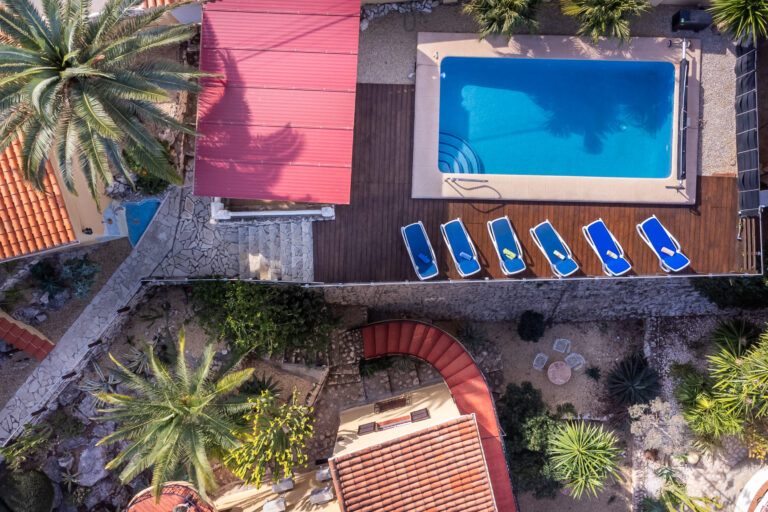 Vista aérea de una piscina rectangular con ocho tumbonas azules, rodeada de palmeras y senderos de piedra.