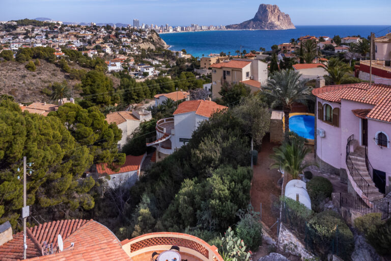 Zona residencial con casas de techo rojo, vegetación, piscina azul y vista al mar con un peñón destacado en el horizonte.