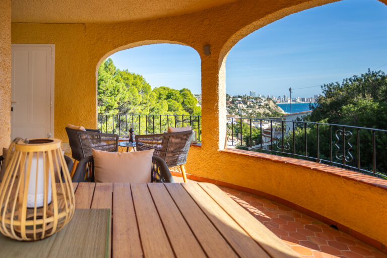Terraza con paredes amarillas, muebles de mimbre y vista al mar, rodeada de árboles y arquitectura urbana a lo lejos.