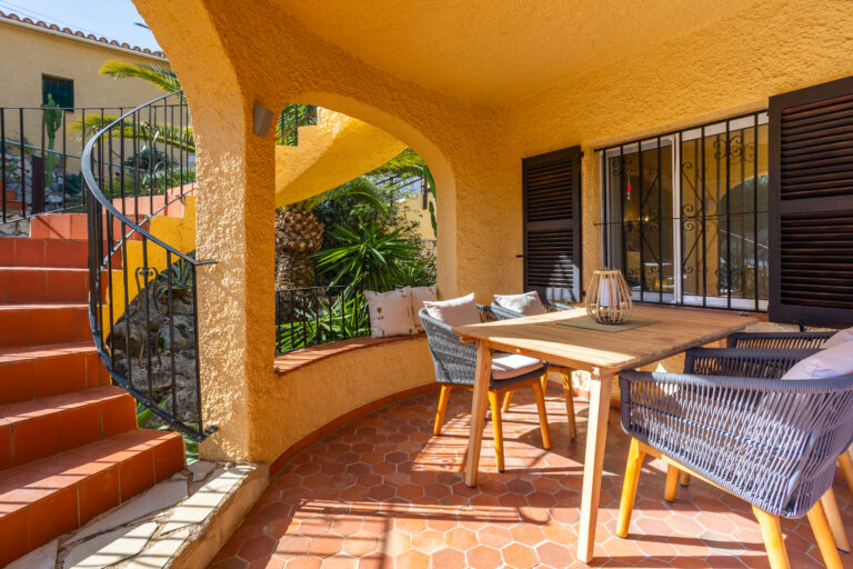 Terraza cubierta con mesa de madera, sillas con cojines, escaleras de ladrillo, plantas y pared amarilla.