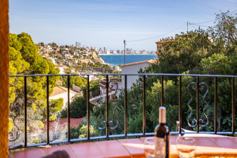 Vista panorámica de ciudad costera desde un balcón con baranda y botella de vino. Edificios, mar azul y vegetación abundante.