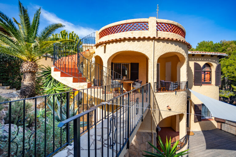Casa de dos niveles con fachada de estuco amarillo, escaleras exteriores curveadas, palmeras y vegetación alrededor.