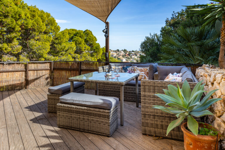 Terraza con mobiliario de ratán, cojines coloridos, mesa de cristal, plantas verdes y vistas a un paisaje urbano verde.