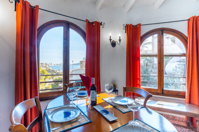 Ventanas arqueadas con cortinas naranjas, mesa de comedor con vajilla elegante y vista al mar.