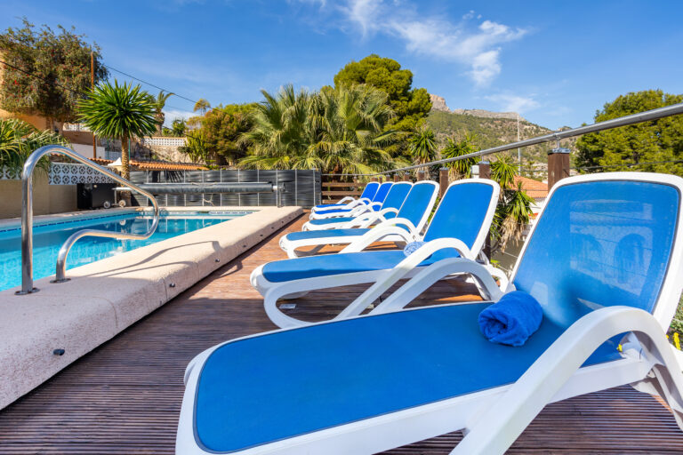Piscina al aire libre con tumbonas azules, rodeada de palmeras y con montañas al fondo bajo un cielo despejado.