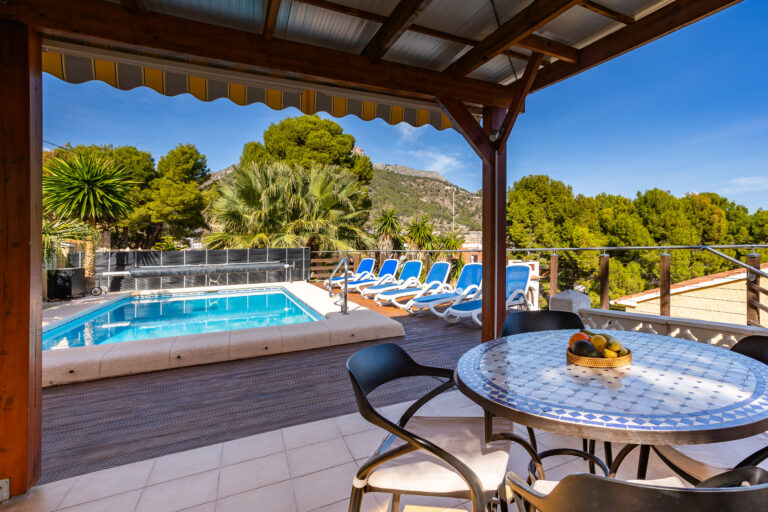 Terraza con piscina, tumbonas azules, mesa redonda con mosaico y sillas; paisaje de montañas y árboles verdes.