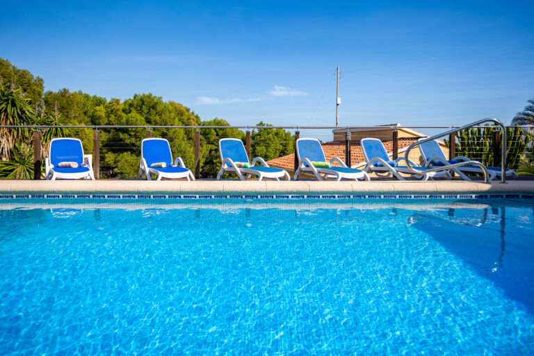 Piscina azul clara con sillas azules alineadas al fondo, cielo despejado y árboles verdes alrededor.