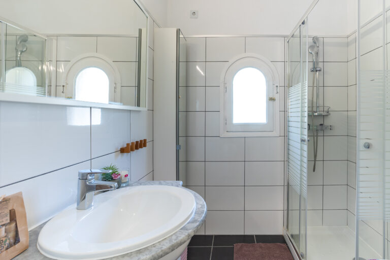 Cuarto de baño moderno con azulejos blancos, ventana arqueada, ducha de cristal, lavabo con grifo plateado y espejo grande.