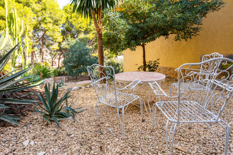 Muebles de jardín de hierro forjado blanco, rodeados de plantas, árboles y grava clara en un entorno soleado.