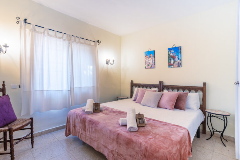 Habitación luminosa con cama doble, edredón rosa, cojines color pastel, cuadros en la pared y luz natural suave.