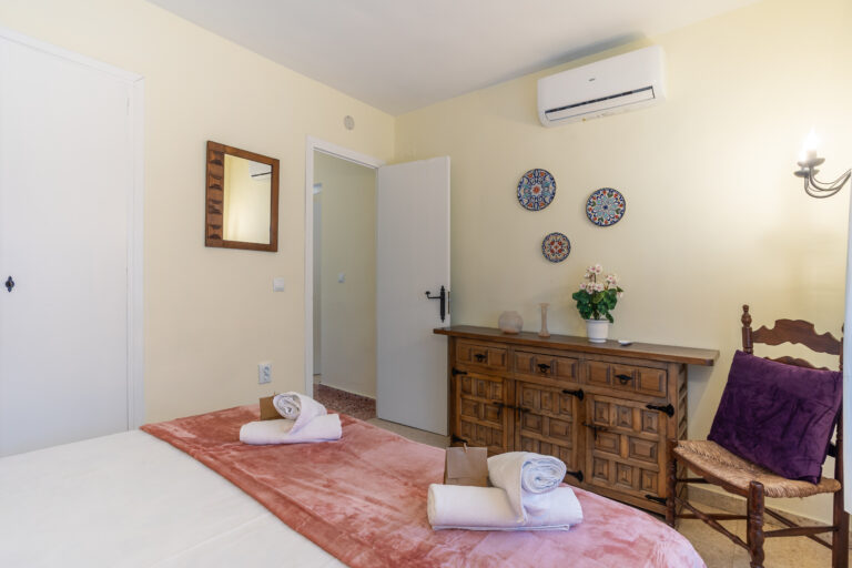 Habitación con paredes claras, cama con edredón rosa, aire acondicionado, cómoda de madera, silla, y tres platos decorativos en la pared.