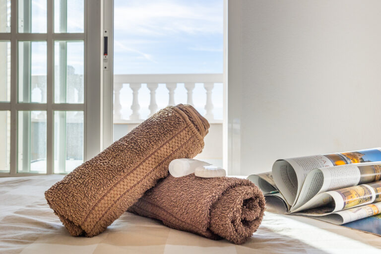 Toallas marrones enrolladas, jabón y botella sobre cama junto a ventana. Revista abierta, fondo de barandilla blanca y cielo azul.