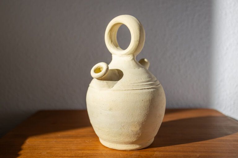 Vasija de cerámica beige con dos picos, cuerpo redondeado y asa circular en la parte superior, sobre una mesa de madera.