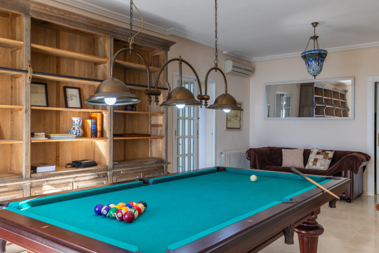 Mesa de billar verde, bolas de colores, lámparas colgantes, estantería de madera, sofá con cojines y luz natural.