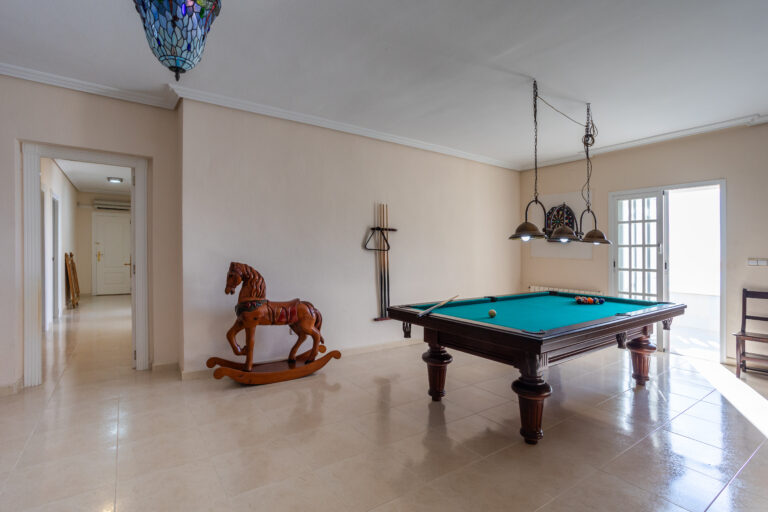 Habitación con mesa de billar, lámpara colgante, caballito de madera, dardos en la pared y puerta hacia un balcón.