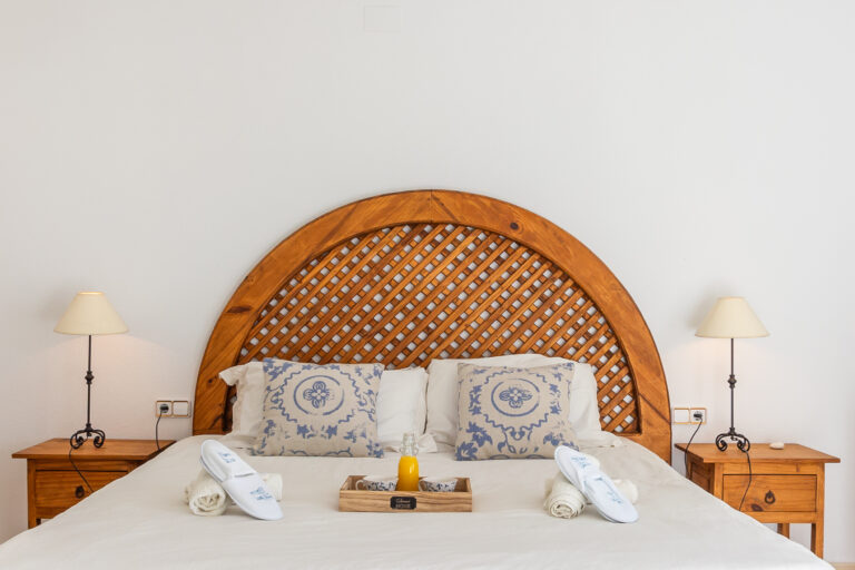 Cama con cabecero de madera trenzada, almohadas decorativas, lámparas en mesas laterales y bandeja con jugo.