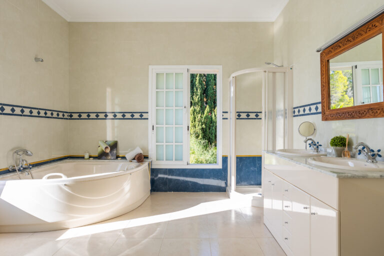 Baño luminoso con bañera ovalada, ventana amplia, ducha, doble lavabo con espejo, y azulejos beige con detalles azules.
