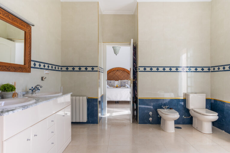 Baño amplio con lavabo doble, espejo con marco de madera, inodoro y bidé. Azulejos beige y azul con mosaicos. Vista a dormitorio.