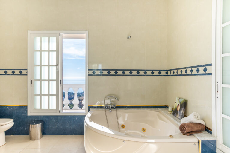 Baño con azulejos beige y azul, bañera de hidromasaje, ventana con vista al mar, toallas enrolladas y papelera cromada.