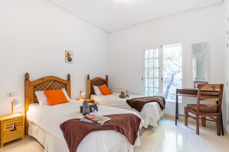 Habitación luminosa con dos camas individuales, cabeceros de madera, cojines y mantas naranjas y marrones, y un escritorio con silla.