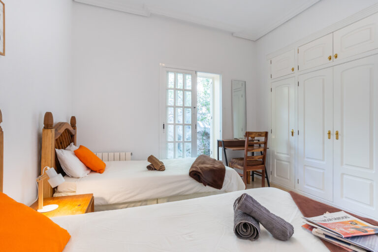 Habitación luminosa con dos camas individuales, cojines naranjas, toallas enrolladas, armario blanco, escritorio y puerta al jardín.