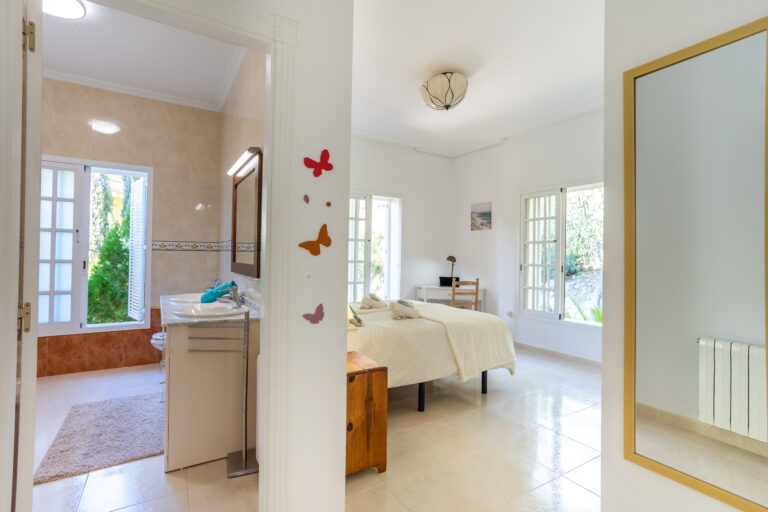 Dormitorio luminoso con paredes blancas, cama doble, ventanas grandes, baño adjunto. Decoración sencilla con mariposas en la pared.