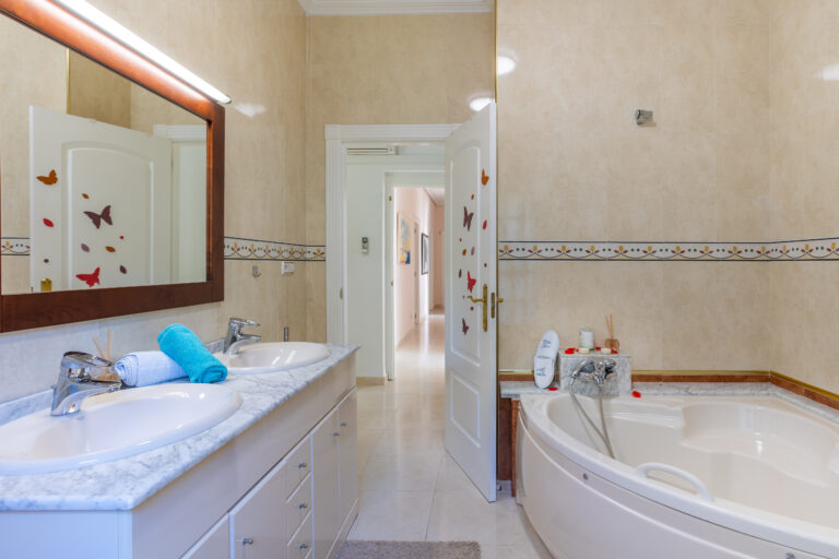 Baño con lavabo doble, encimera de mármol, bañera esquinera, espejo grande y toallas coloridas. Decoración de mariposas en la puerta.