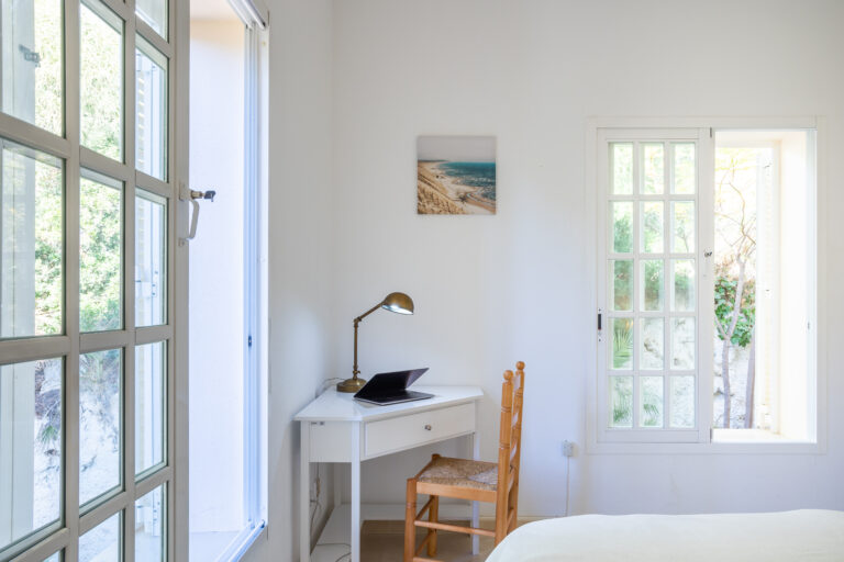 Habitación luminosa con escritorio blanco, silla de madera y lámpara. Ventanas grandes, paisaje exterior verde.