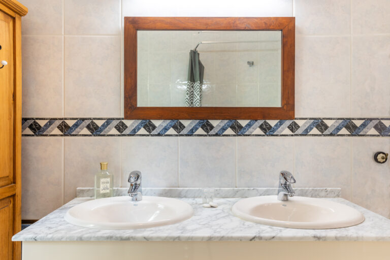 Baño con dos lavabos blancos, mármol, espejo de marco de madera, azulejos decorativos y botella transparente.