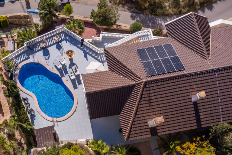 Tejado con paneles solares, piscina en forma de riñón con agua azul, terraza con sillones y sombrilla blanca, vegetación alrededor.