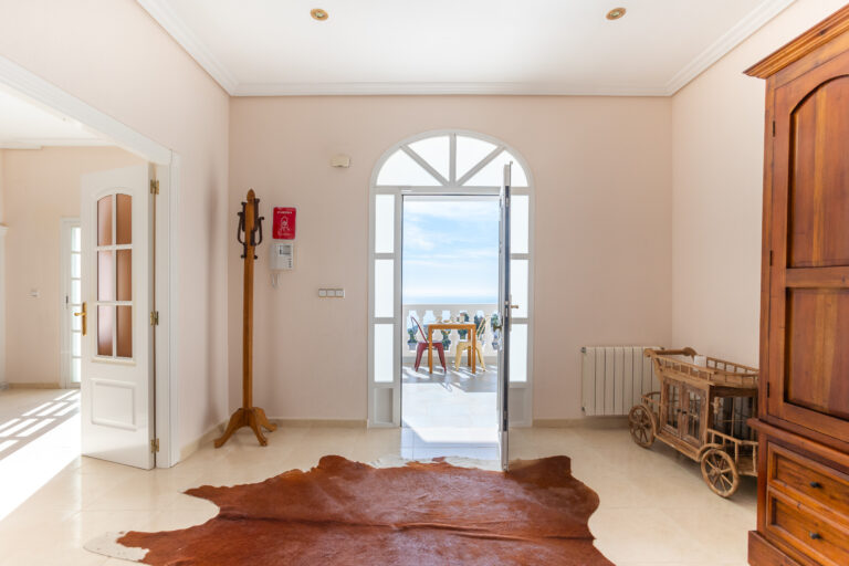 Vestíbulo luminoso con puerta abierta a balcón, vista al mar, alfombra piel, armario de madera y carrito decorativo.