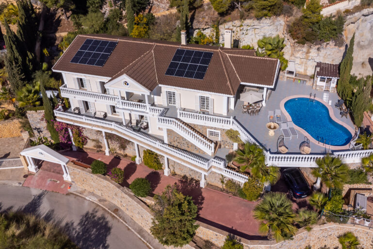 Casa blanca con paneles solares, piscina azul y amplio patio. Rodeada de vegetación en una colina. Camino de acceso curvado.