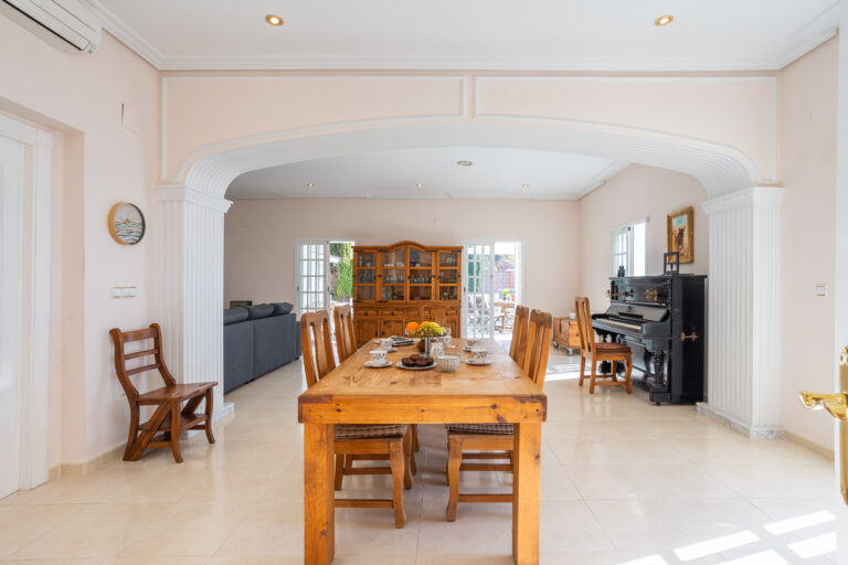 Comedor con mesa de madera, siete sillas, piano negro, sofá gris y vitrina. Paredes claras y piso beige brillante.