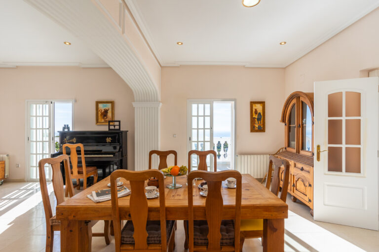 Comedor luminoso con mesa de madera, sillas, piano negro, muebles clásicos y obras de arte; vista al exterior con balcón.