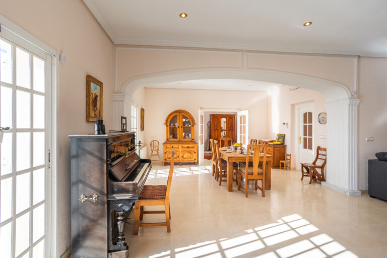 Sala luminosa con piano vertical negro, mesa de madera con sillas, vitrina y puertas de cristal, decorada con suaves tonos pastel.