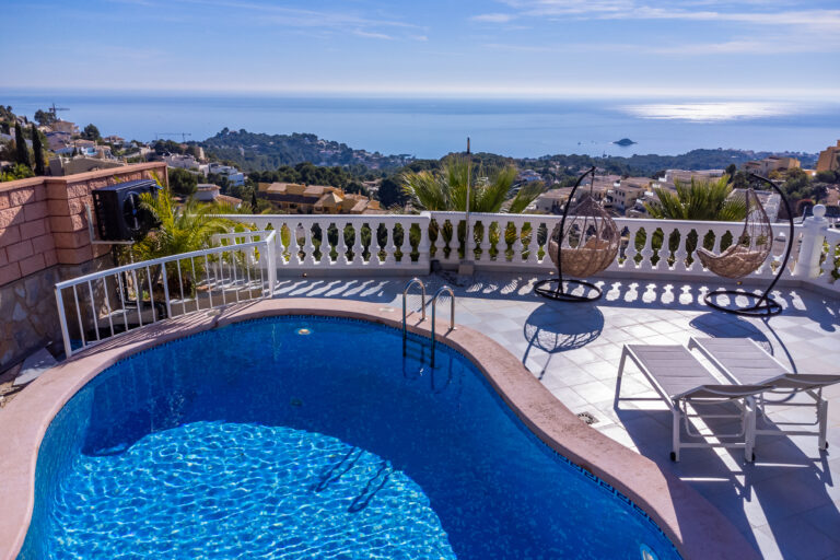 Piscina azul en terraza con vista al mar, sillas colgantes, tumbonas blancas y paisaje de colinas y casas bajo un cielo despejado.
