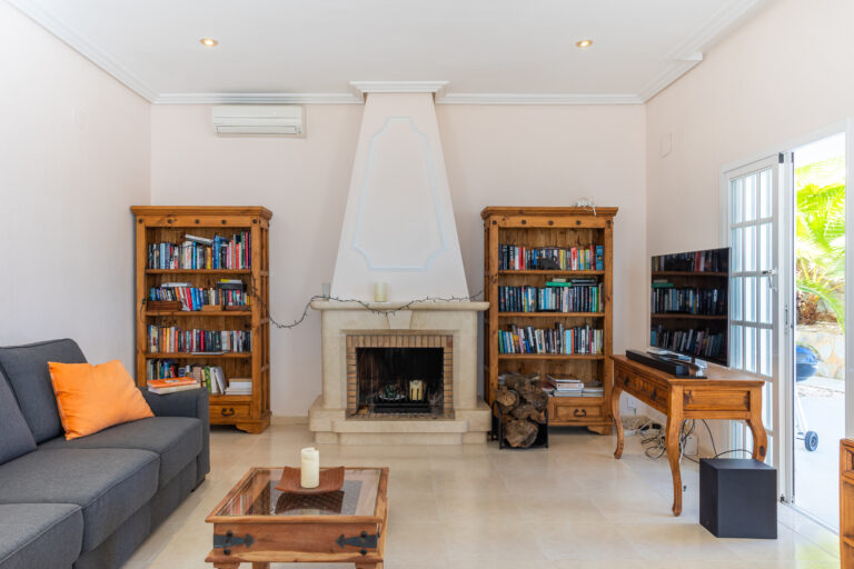Sala de estar con sofá gris, cojín naranja, dos estanterías llenas de libros, chimenea, mesa de centro y televisor cerca de la ventana.