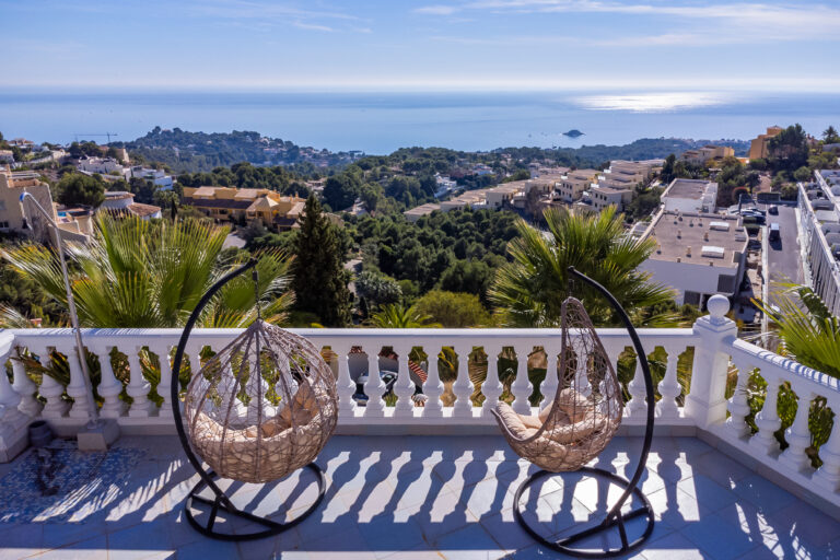 Terraza con dos sillones colgantes, vista al mar y colinas, rodeada de palmeras y edificios en un día soleado.