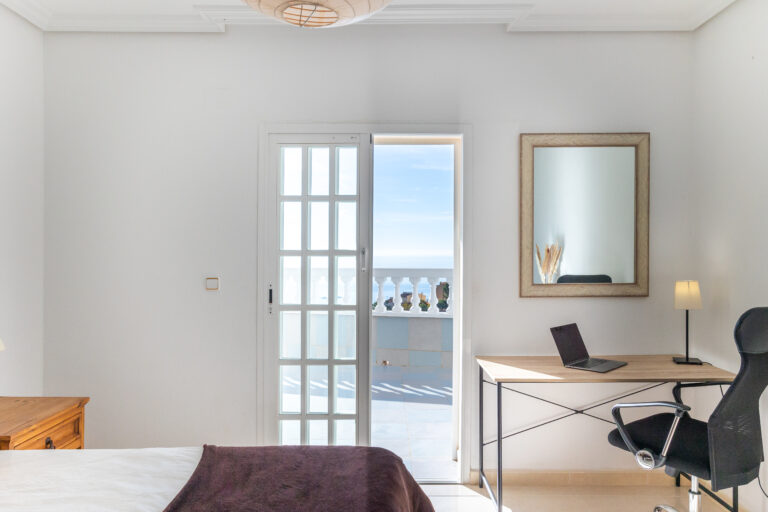 Habitación luminosa con escritorio, ordenador portátil, silla ergonómica, lámpara y puerta a balcón con vista al mar.