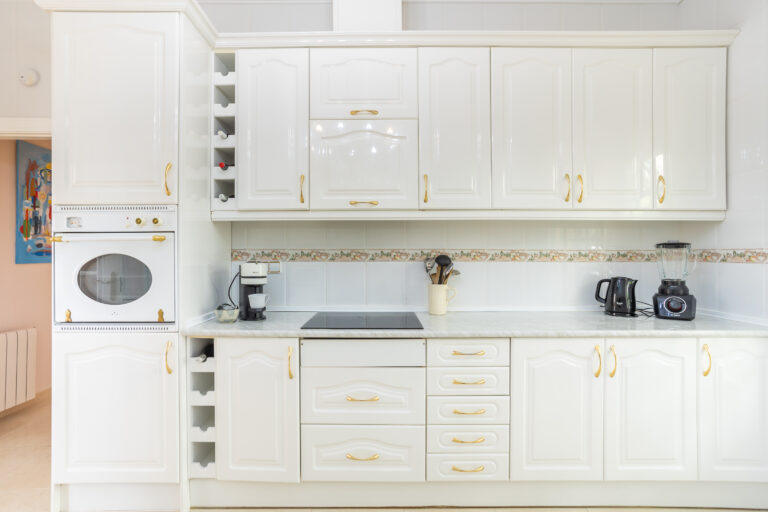 Cocina blanca con gabinetes y tiradores dorados, horno empotrado, estufa, cafetera, licuadora y tetera sobre encimera.
